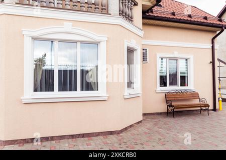 Die Fassade eines Hauses mit großen Fenstern mit weißen Rahmen, beigefarbenen Wänden und einem rot gekachelten Dach. Eine hölzerne Bank mit schmiedeeisernen Details ist Plac Stockfoto