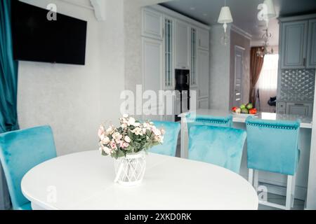 Moderner Essbereich mit blauen Samtstühlen, einem weißen Tisch, einem floralen Herzstück und Blick auf eine stilvolle Küche. Stockfoto