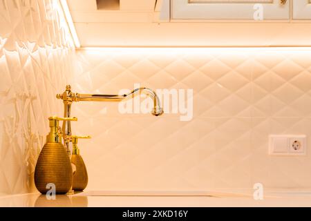 Nahaufnahme einer goldenen Küchenarmatur und Seifenspender gegen eine weiße Rautenfliese. Warme Beleuchtung unterstreicht das elegante Design. Stockfoto