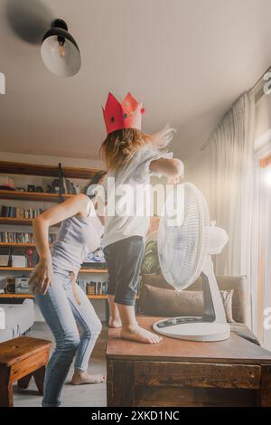 Mutter und Tochter in Kronen tanzen fröhlich vor einem Ventilator an einem heißen Sommertag zu Hause, genießen familiäre Zweisamkeit und unbeschwerte Freude Stockfoto