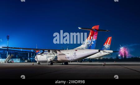 Ein Flugzeug mit weißem Körper und rotem Schwanz parkt nachts auf einem Asphalt. Hinter dem Flugzeug explodieren Feuerwerke. Stockfoto