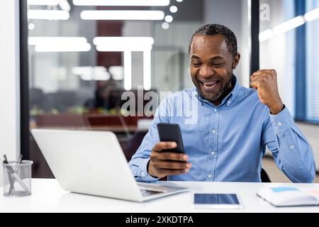 Glücklicher Geschäftsmann, der Erfolg mit Smartphone bei der Arbeit feiert. Fröhlicher Büroangestellter zeigt Aufregung, blickt auf den mobilen Bildschirm und macht Faustpumpen. Arbeitsplatz mit Laptop Stockfoto