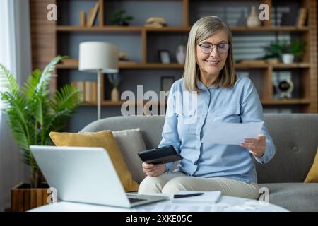 Ältere Frau sitzt auf der Couch, benutzt Laptop und überprüft Finanzdokumente. Konzept der Eigenheimfinanzen, Budgetverwaltung und Senior Independent liv Stockfoto