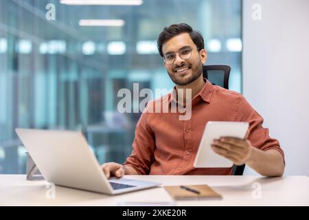 Indischer Programmierer, der in einem modernen Büro mit Laptop und Tablet arbeitet. Professionelles Lächeln, Einsatz von Technologie für Aufgaben, Produktivität. Moderner Arbeitsbereich, Glaswände, technische Geräte. Stockfoto