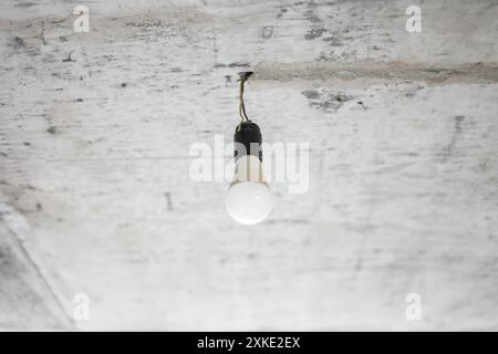 Weiße Glühlampe mit Drähten oder Kabeln an rauer, grauer Betondecke im Innenbereich in unvollendeter Renovierung. Durchführung von Reparaturen, Verbesserung der Wohnbedingungen. Alte Industrie, Kellerraum, Bombenschutzraum Stockfoto