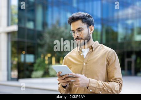 Junger Mann, der vor einem modernen Glasbürogebäude mit einem digitalen Tablet steht. Konzentration auf arbeitsbezogene Aufgaben, die einen professionellen und technisch versierten Lebensstil in der Stadt widerspiegeln. Stockfoto