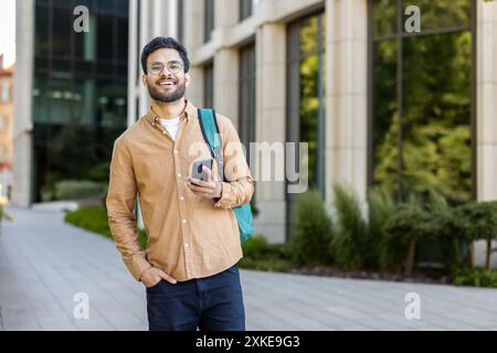 Selbstbewusster junger indischer Student, der ein Smartphone hält, lächelt und draußen auf einem sonnigen Universitätscampus steht. Das Konzept der Bildung, Jugend und Technologie. Stockfoto