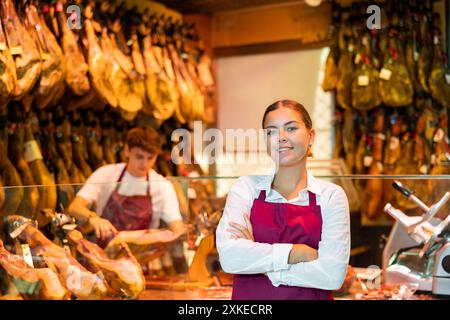 Junge weibliche Metzgerin mit gekreuzten Armen in der Nähe hängender Beine mit iberischem Schinken Stockfoto