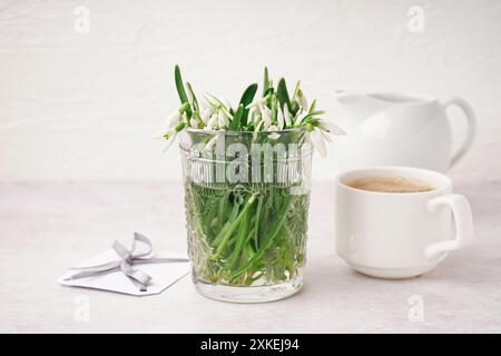 Glas mit wunderschönen Schneetropfen, Postkarte und Tasse Kaffee auf grauem Grunge-Hintergrund Stockfoto