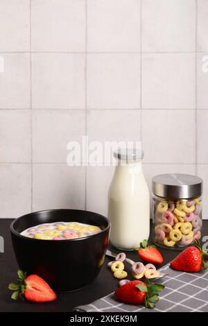 Schüssel mit bunten süßen Müsliringen mit Milch und Erdbeeren auf schwarzem Tisch Stockfoto