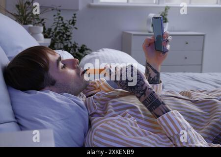 Junger Mann im Pyjama, der elektronische Einweg-Zigarette raucht und das Handy zu Hause auf dem Bett benutzt Stockfoto