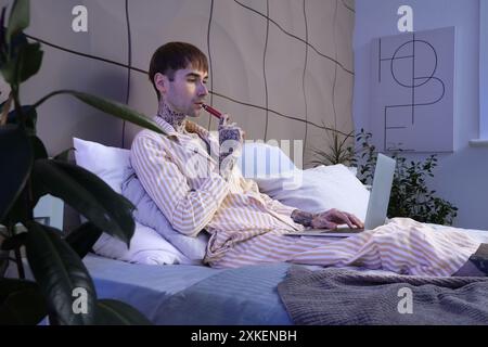 Junger Mann im Schlafanzug mit elektronischer Einwegzigarette mit Laptop auf dem Bett zu Hause Stockfoto