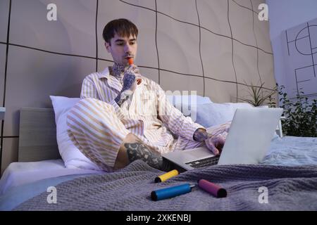 Junger gutaussehender Mann im Pyjama, der verschiedene elektronische Einweg-Zigaretten raucht und Laptop auf dem Bett zu Hause benutzt Stockfoto