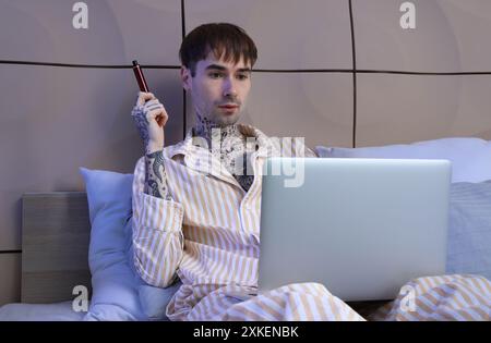 Junger Mann im Schlafanzug mit elektronischer Einwegzigarette mit Laptop auf dem Bett zu Hause Stockfoto