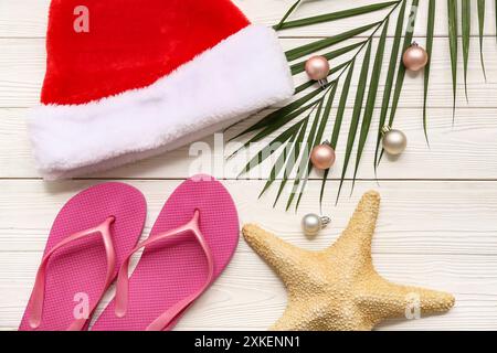 Flipflops, Seesterne, Weihnachtsmannmütze und Weihnachtskugeln auf Palmenzweig auf weißem Holzhintergrund. Draufsicht Stockfoto