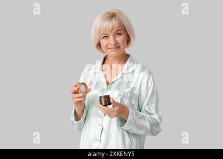 Schöne reife glückliche Frau im Pyjama mit Glas Gesichtscreme auf grauem Hintergrund Stockfoto