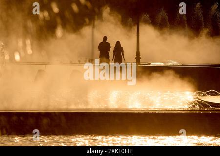 Bukarest, Rumänien. 22. Juli 2024: Menschen laufen an einem sehr heißen Tag durch den feinen Sprühnebel eines Brunnens. Quelle: Lucian Alecu/Alamy Live News Stockfoto