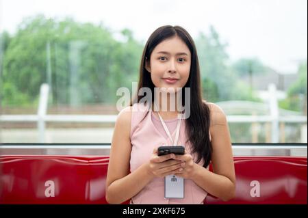 Eine positive junge asiatische Büroangestellte mit einem Smartphone in der Hand lächelt in die Kamera, während sie auf einem Sitz in einem Himmelszug sitzt und pendelt Stockfoto