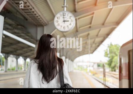 Ein Rückansicht einer asiatischen Frau mit einem Rucksack, die auf einer Plattform steht, auf eine Uhr blickt oder ihre Ankunftszeit mit öffentlichen Verkehrsmitteln überprüft Stockfoto