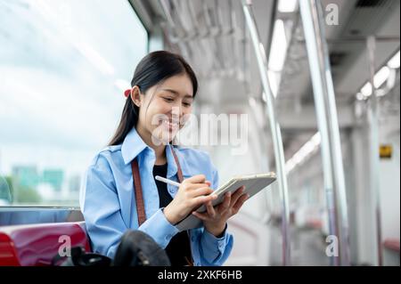 Eine glückliche, schöne junge asiatische Passagierin, die ihr digitales Tablet in einem Skytrain benutzt und mit öffentlichen Verkehrsmitteln in der Stadt pendelt. Lifestyle, trave Stockfoto