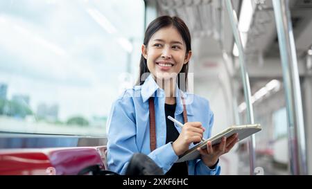 Eine schöne junge asiatische Passagierin träumt tagträumen und blickt auf den Blick nach draußen, während sie ihr digitales Tablet in einem Himmelszug benutzt und im C pendelt Stockfoto