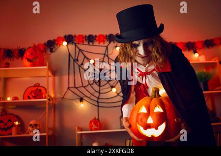 Gruseliges Porträt eines Vampirmannes, der im Dunkeln im Zimmer steht, mit leuchtender Jack-o-Laterne in den Händen. Stockfoto