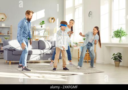Große, fröhliche Familie spielt Spiele mit verbundenen Augen im Wohnzimmer. Kinder und Eltern nehmen an zeitlosen Spielen des Blinden-Mannes-Buff-Spiels Teil, das Licht bringt Stockfoto