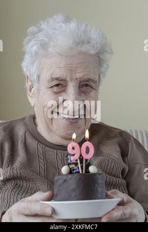 Eine hübsche und glückliche Seniorin, die neunzig Geburtstagskuchen mit Kerzen hält Stockfoto