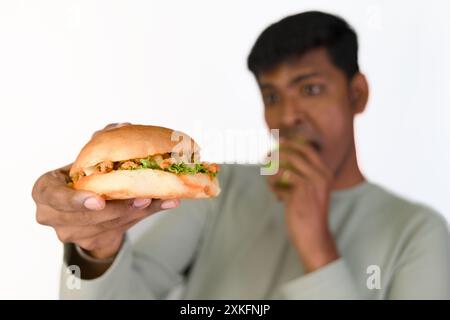 Ein Mann kann sich nicht entscheiden, was er zuerst verschlingen soll, indem er einen saftigen Burger hält, während er einen Hauch von Apfel nimmt. Stockfoto