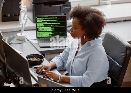 Hochwinkel-Seitenansicht eines jungen afroamerikanischen Softwareentwicklers, der Code am Computer schreibt, während er am Schreibtisch sitzt, mit einer Schüssel zur Hand und mehreren Bildschirmen in einem modernen Büro Stockfoto