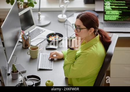 Seitenansicht einer jungen rothaarigen Softwareingenieurin mit grünem Hemd und einer Brille, die sich auf das Lesen des Computerbildschirms konzentriert, während sie am Schreibtisch im modernen Programmierbüro sitzt Stockfoto