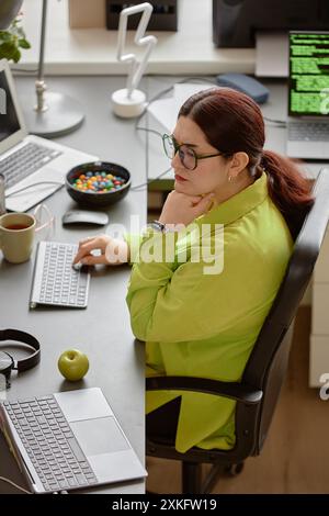 Vertikale Aufnahme einer durchdachten IT-Fachfrau, die in modernen Büros Computerbildschirm liest und auf der Tastatur ein Skript eingibt Stockfoto