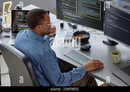 Hochwinkel-Seitenansicht eines nachdenklichen männlichen afroamerikanischen Softwareentwicklers, der Computerbildschirm liest und Fehler im Code findet, während er am Schreibtisch sitzt, mit Tee- und Süßigkeitenschüssel zur Hand im Büro, Kopierraum Stockfoto