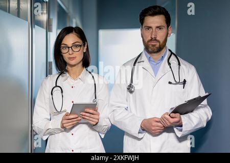 Zwei Fachärzte stehen im Krankenhausflur. Weibliche Ärztin mit Tablette. Männlicher Arzt mit Klemmbrett. Fachärzte in formaler Kleidung mit weißen Mänteln und Stethoskopen. Stockfoto