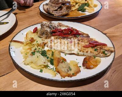 Weiße Platte mit gegrilltem Wolfsbarsch, roter Paprika und Kartoffelstapeln oder gebratenen Kartoffelscheiben auf einem Holztisch in einem Restaurant oder zu Hause Stockfoto