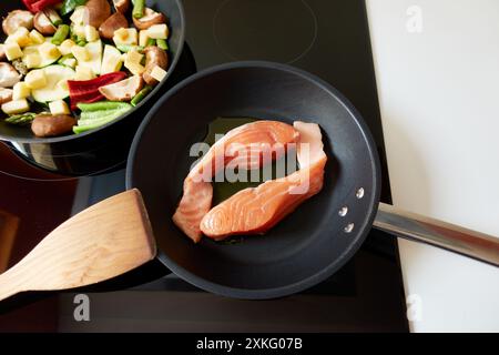 Zwei Pfannen kochen auf dem Induktionsherd: Eine mit zwei Stücken Lachsfischlende und die andere mit geschnittenem Gemüse wie grünem und rotem Pfeffer, zucc Stockfoto