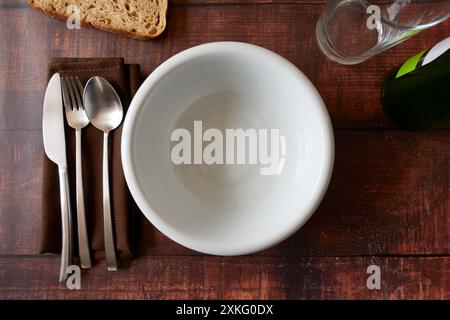 Blick von oben auf einen leeren weißen tiefen Teller mit Löffel, Gabel und Messer, Stoffserviette, Glasbecher und Flasche auf Holztisch Stockfoto