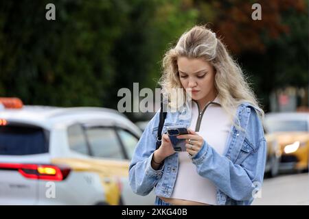 Attraktives Mädchen mit blondem lockigem Haar, das mit Smartphone in den Händen auf einer Straße auf Taxi-Autos-Hintergrund steht Stockfoto