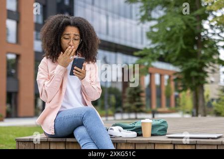 Eine junge Frau mit lockigen Haaren und Brille sieht schockiert auf ihr Smartphone aus, während sie auf einer Bank in einem Stadtgebiet sitzt. Rucksack, Kaffeetasse und Laptop neben ihr. Stockfoto