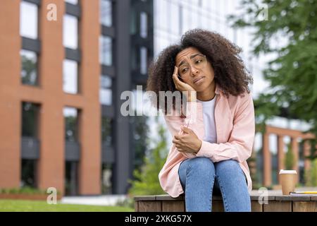 Nachdenkliche junge Frau mit lockigem Haar, die draußen in städtischer Umgebung sitzt. Sie wirkt nachdenklich und besinnlich. Brillen sorgen für einen intellektuellen Look Der moderne Gebäudehintergrund lässt urbanen Lebensstil vermuten Stockfoto