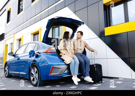 Junge Paare, Mann und Frau, die mit einem Elektroauto unterwegs sind, halten an der Ladestation. Freund und Freundin sitzen im Kofferraum und lächeln. Stockfoto