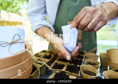 Gärtner, der Samen in biologisch abbaubare Töpfe pflanzt. Hände mit Erde, Töpfen und Samenpaketen. Gartenarbeit an einem sonnigen Tag. Stockfoto