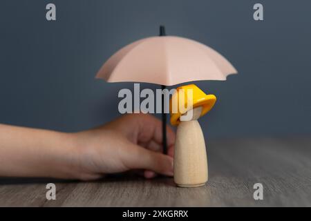 Nahaufnahme einer Hand, die einen Regenschirm über einer Holzpuppe hält und einen gelben Schutzhelm trägt. Mitarbeiter- und Mitarbeiterversicherungskonzept. Stockfoto