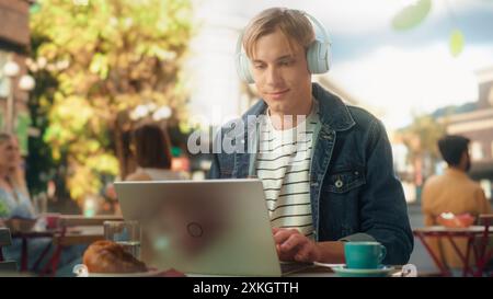 Junger, stilvoller freiberuflicher Designer, der ein Online-Gespräch mit einem Kunden führt. Content Creator in Kopfhörern mit Laptop-Computer, um von einer ruhigen City Restaurant Summer Terrace aus ferngesteuert zu arbeiten Stockfoto