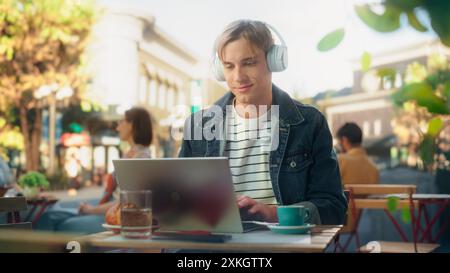 Freiberuflicher Mitarbeiter mit weißen Kopfhörern und einem Laptop für Remote-Online-Arbeiten in der Stadt. Junger kreativer Mann beim Frühstück und Telearbeit von einem Café an einem sonnigen Sommertag Stockfoto