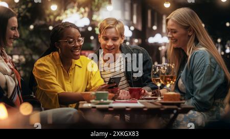 Junge Freunde, die gemeinsam lustige Gespräche in einem Café führen. Stilvolle afrikanische Frau sitzt auf einer Nachterrasse mit multiethnischen Freunden und zeigt lustige Social Network Posts und Nachrichten auf einem Smartphone Stockfoto