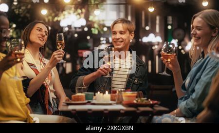 Verschiedene Freunde, die das Nachtleben in einem Street Cafe genießen. Junge Frauen und Männer sitzen hinter einem Tisch, haben lustige und freudige Gespräche. Kaukasischer Mann Teilt Unterhaltsame Lebensgeschichten Stockfoto