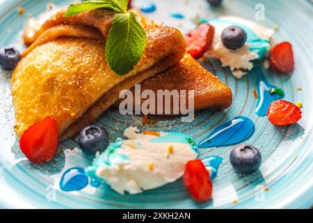 Pfannkuchen oder Crepe suzette mit Quark Creme, Erdbeeren, Heidelbeeren mit Blue Curacao Sauce und Orangenschale auf einem türkisfarbenen Teller. Nahaufnahme. Ausgewählt Stockfoto