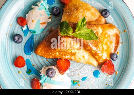 Pfannkuchen oder Crepe suzette mit Quark Creme, Erdbeeren, Heidelbeeren mit Blue Curacao Sauce und Orangenschale auf einem türkisfarbenen Teller. Nahaufnahme. Ausgewählt Stockfoto