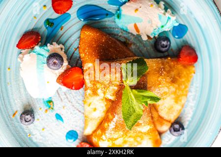 Pfannkuchen oder Crepe suzette mit Quark Creme, Erdbeeren, Heidelbeeren mit Blue Curacao Sauce und Orangenschale auf einem türkisfarbenen Teller. Nahaufnahme. Ausgewählt Stockfoto